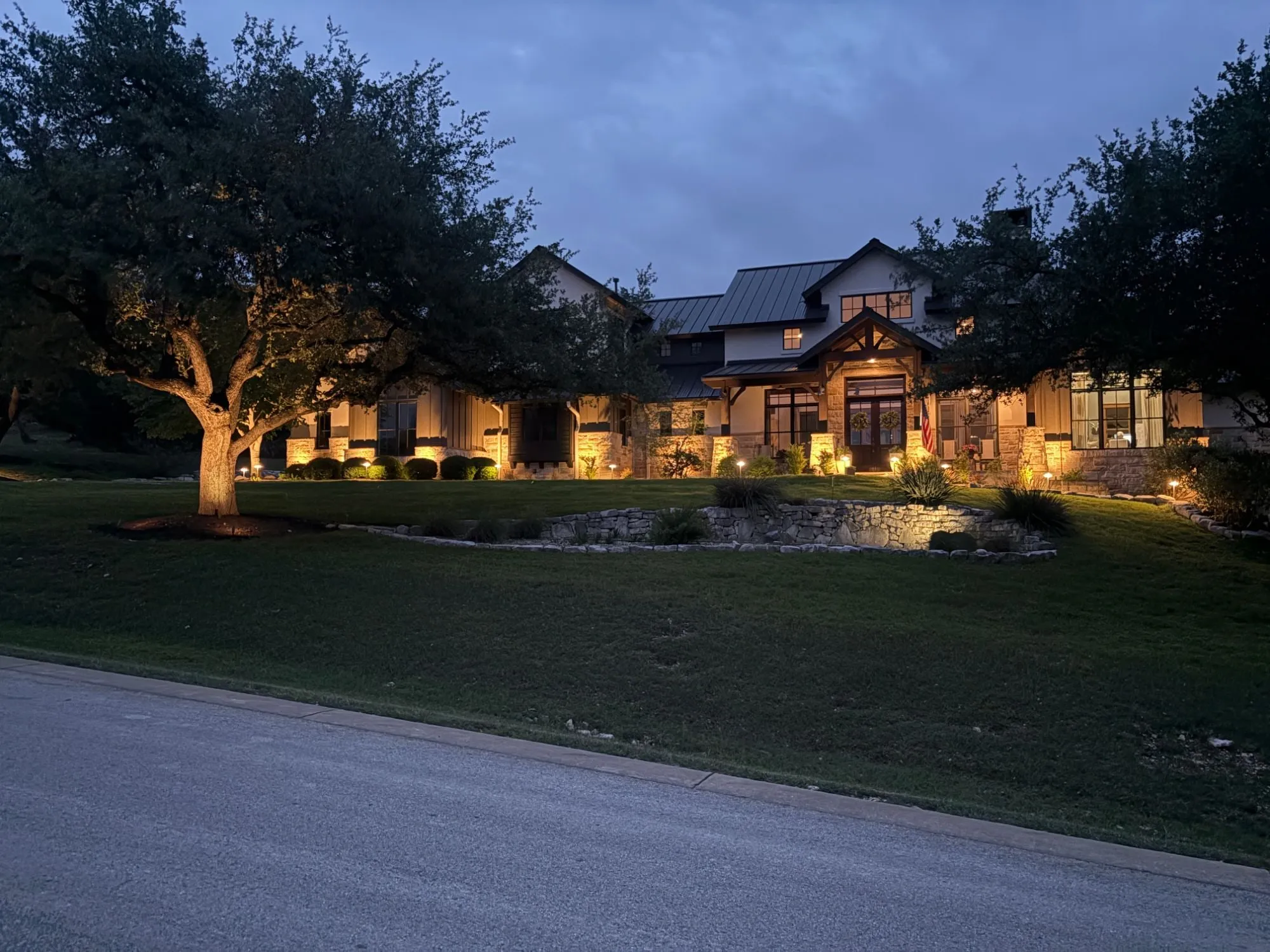 Hill Country estate seen from the street, with lit stone columns, an uplit live oak, path lights, and a glowing stone retaining wall