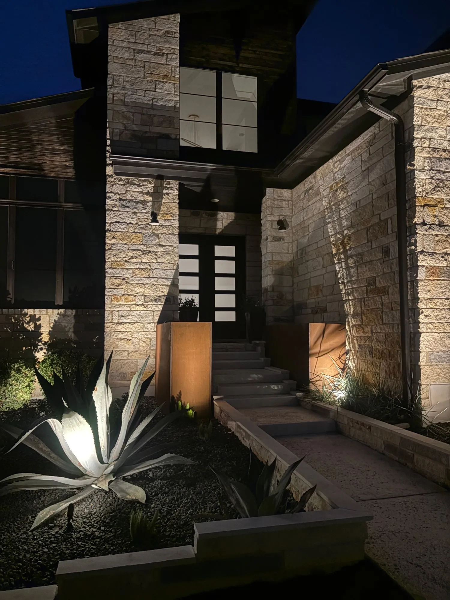 Modern stone entry with an uplit agave, steel planters, and grazed stone walls