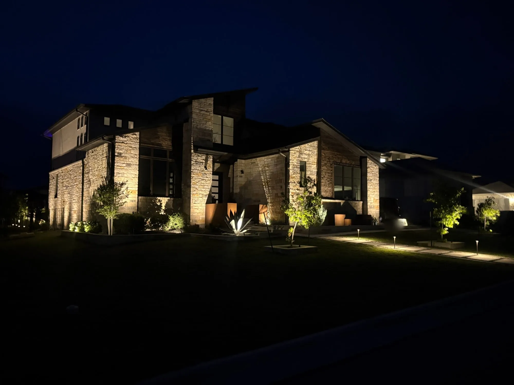 Contemporary stone home grazed with light from end to end, with uplit trees, an uplit agave, and path lights