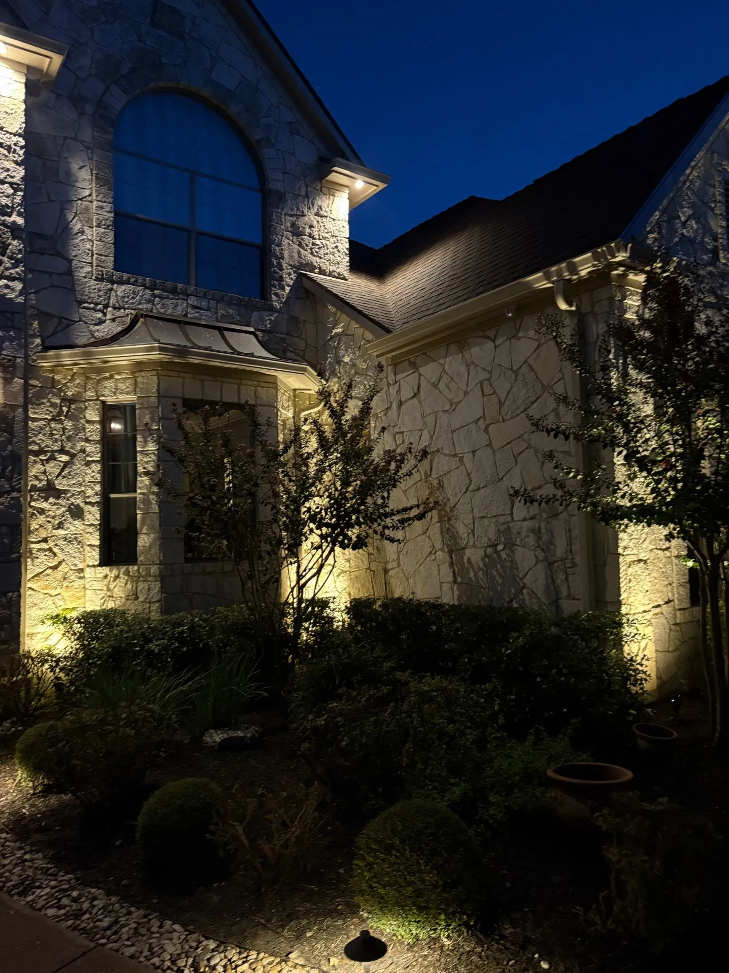 Stone facade with an arched window, softly grazed with uplights between crepe myrtles