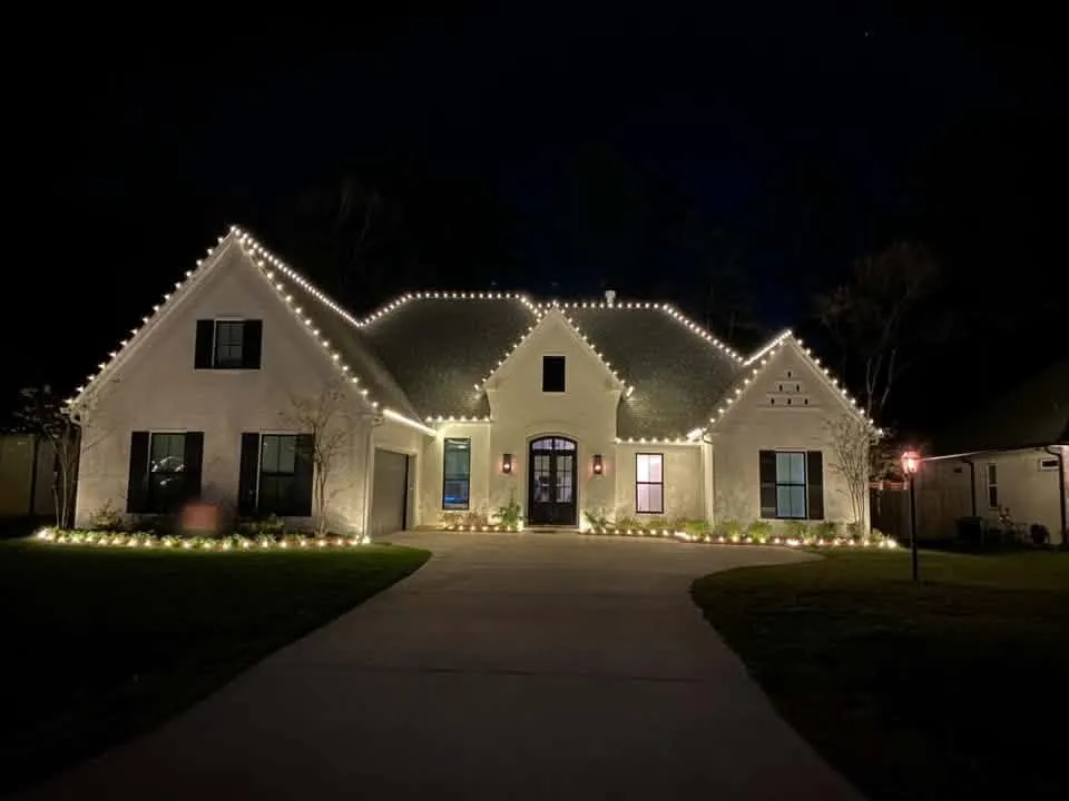 Classic warm white bulbs tracing every roofline and gable of a white brick home, with lit flower beds