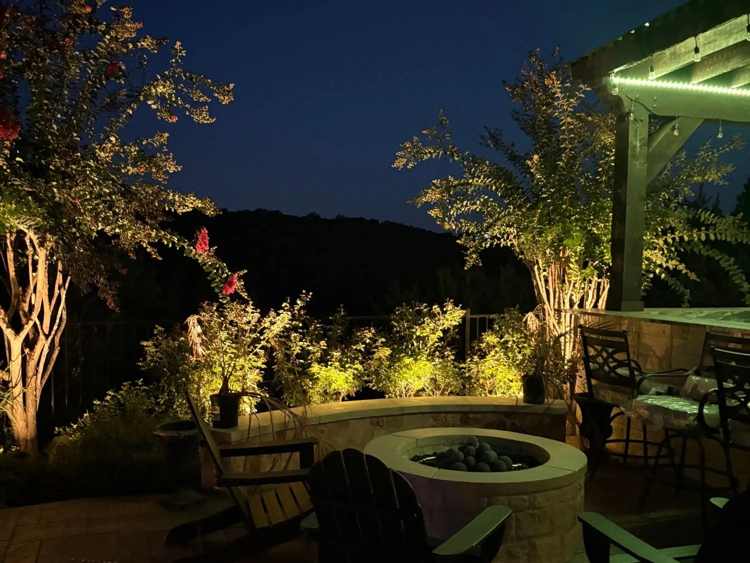 Backyard patio with fire pit, lit seat wall planters, and uplit crepe myrtles overlooking the Hill Country