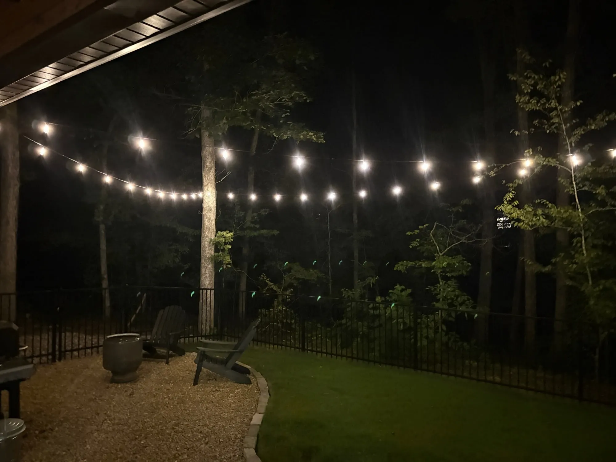 Café string lights over a backyard seating area with a fire pit, lawn, and wooded view