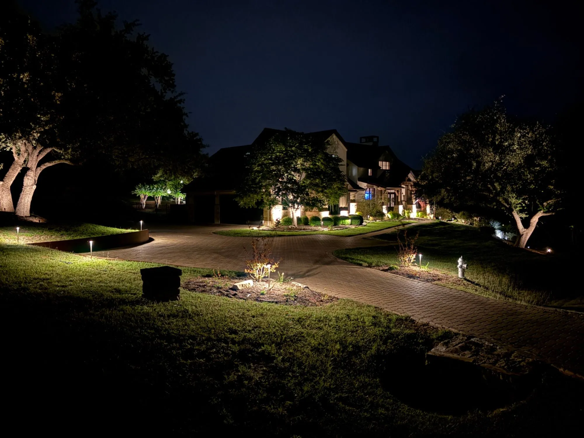 Long paver driveway lined with path lights and uplit oaks